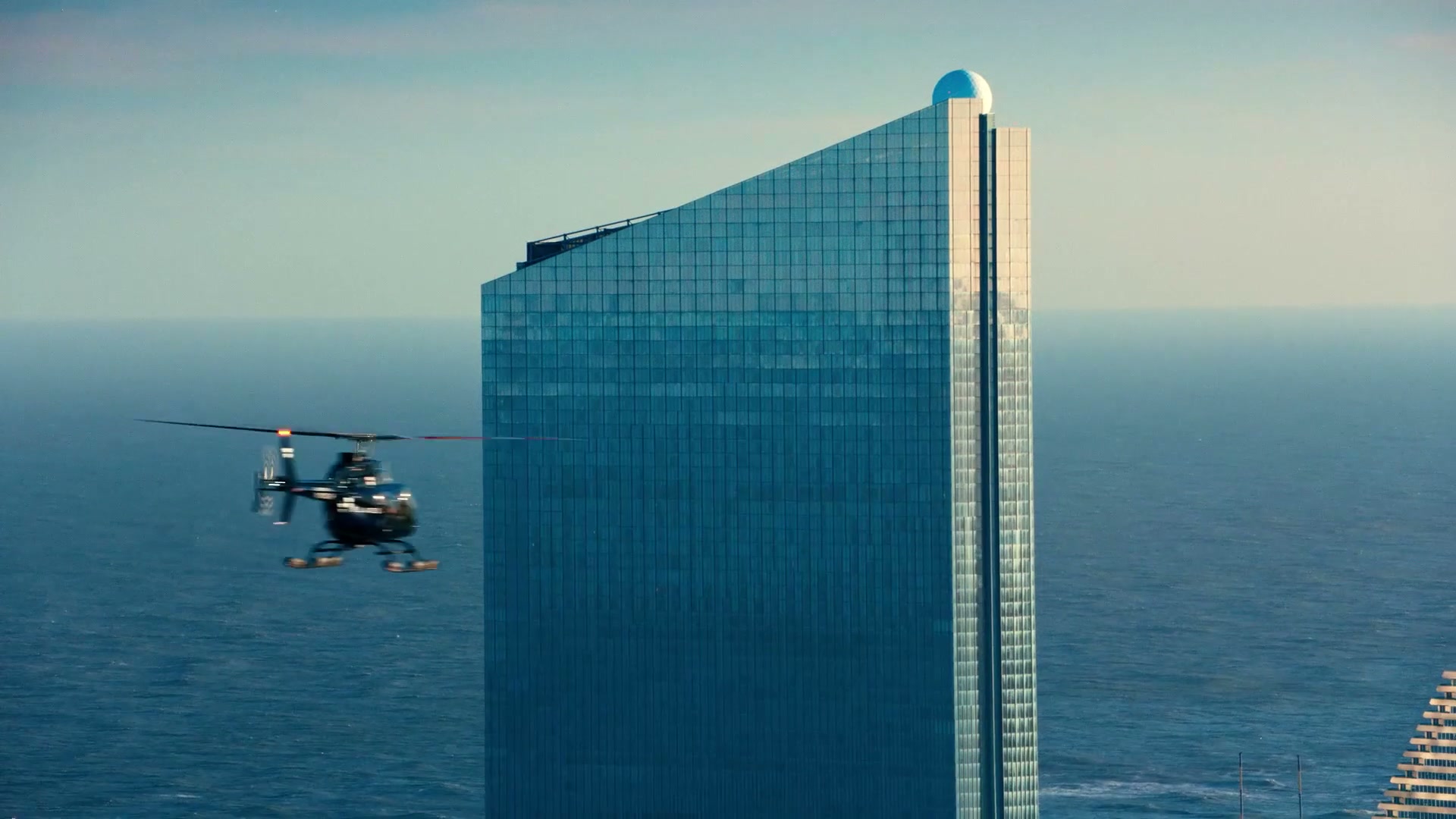 A helicopter sweeping past a glass tower over the ocean
