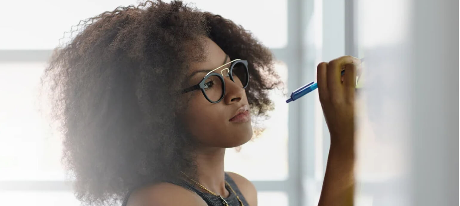 A woman in glasses writing on a glass board, pausing in thought.