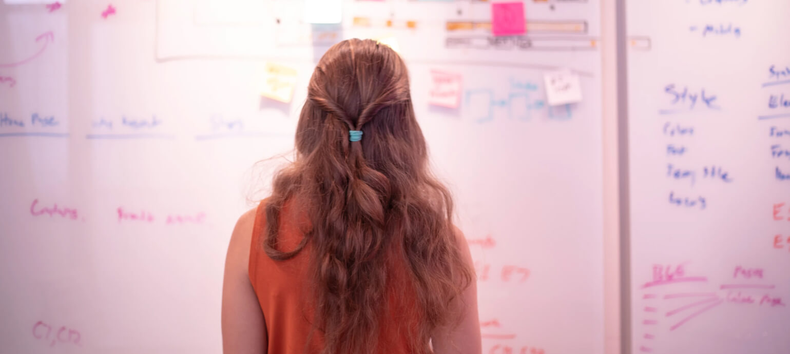 A woman seen from behind studying a whiteboard covered in brand strategy notes, diagrams, and sticky notes.